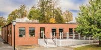 A modern permanent modular building with a red brick exterior and accessible entrance surrounded by trees.